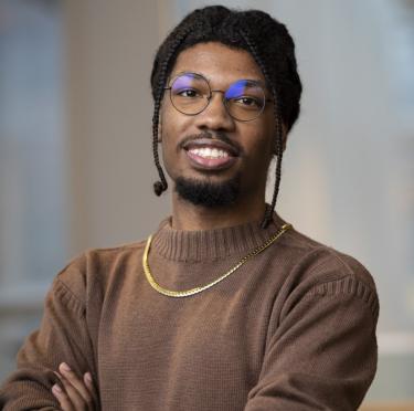 Clifmon Leroy has his arms crossed smiling at the camera while wearing a brown sweater and gold chain