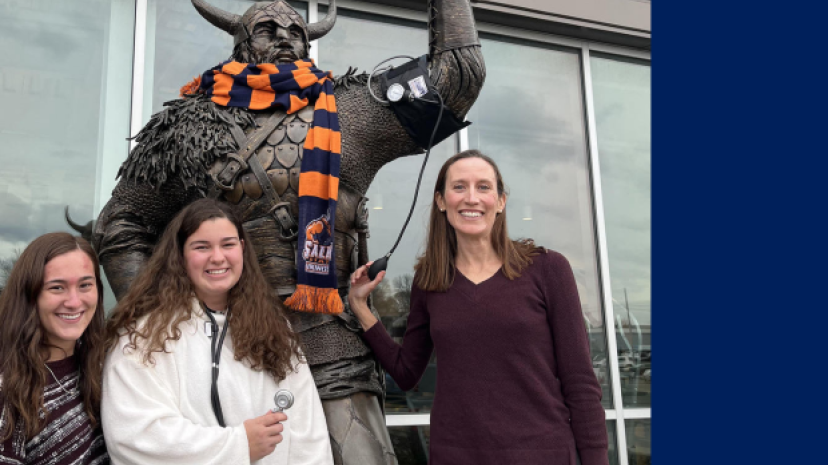 Brett Ely with students in front of the Viking statue, which has an orange and blue Salem State scarf around its neck. One student holds a stethoscope and Professor Ely holds a blood pressure device.