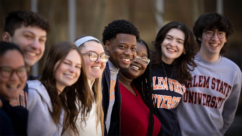 A diverse group of Salem State University students stand closely together outdoors, smiling and many wearing Salem State apparel, representing campus unity and belonging.
