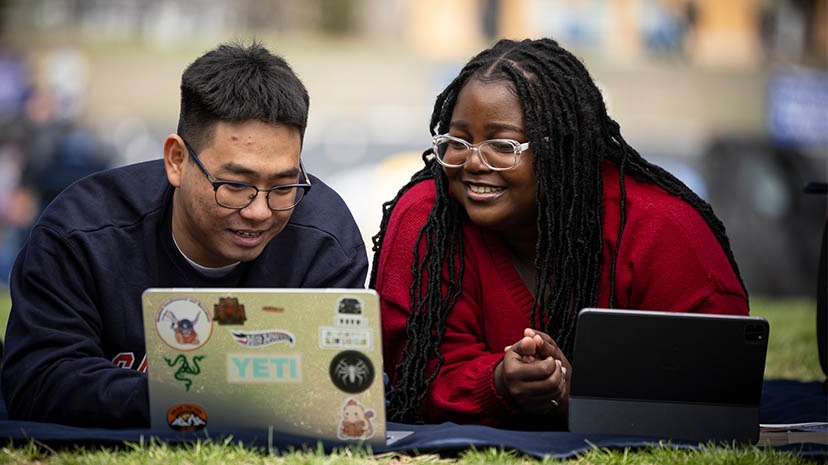 Two Salem State University students lie on a blanket outdoors, smiling as they work together on a laptop and tablet.