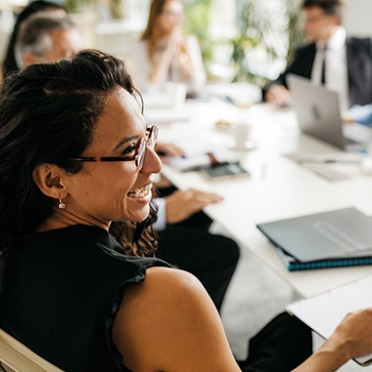 A woman in glasses laughing with colleagues around a table